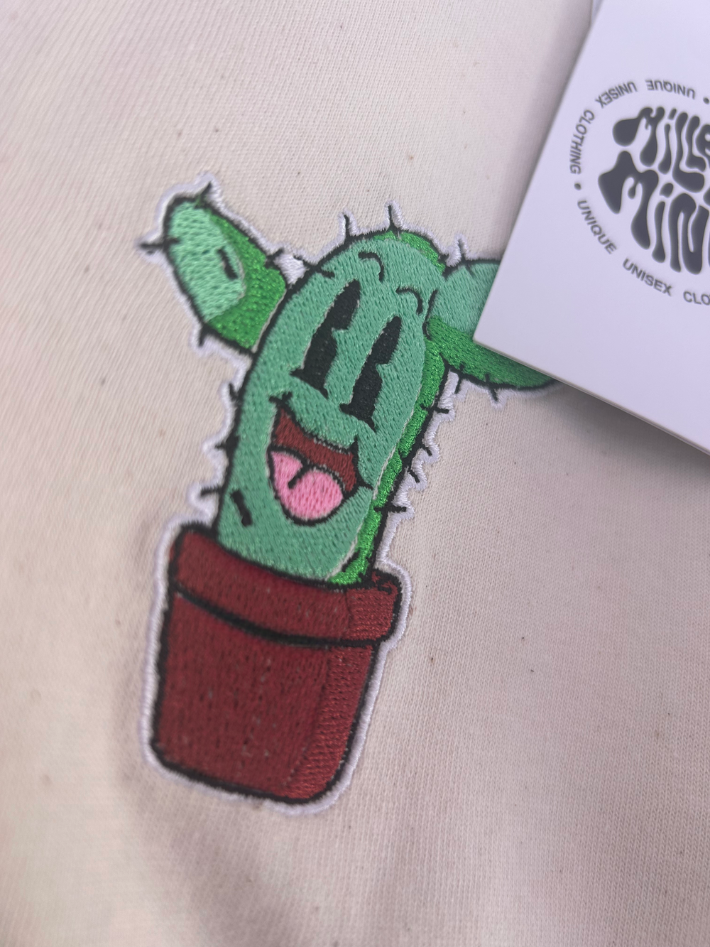 Close-up of a green cartoon cactus patch in a red pot on a light beige fabric, with part of a white printed tag visible at the top right.