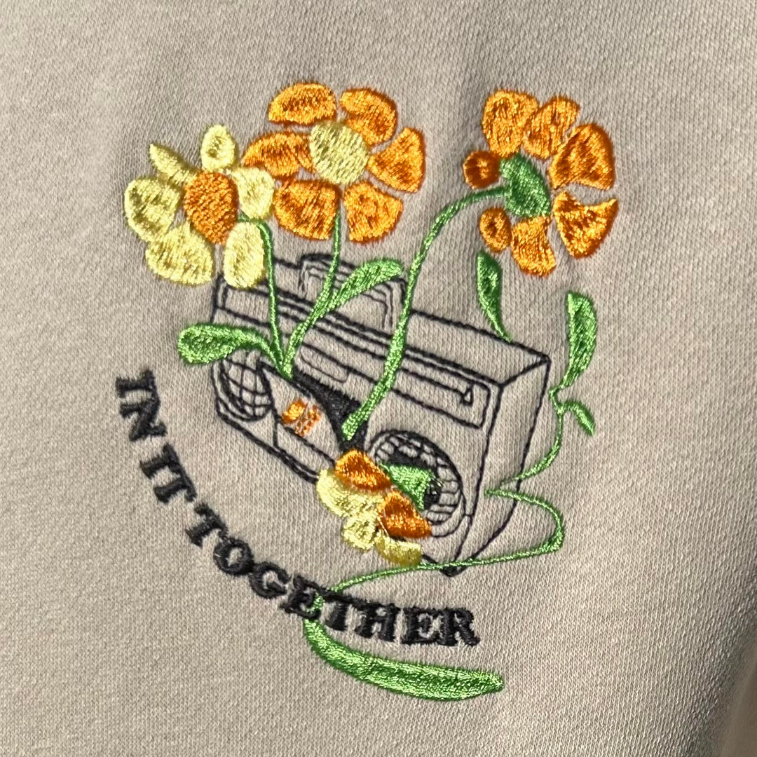 Embroidered design of a boombox with flowers and 'IN IT TOGETHER' text on a beige background