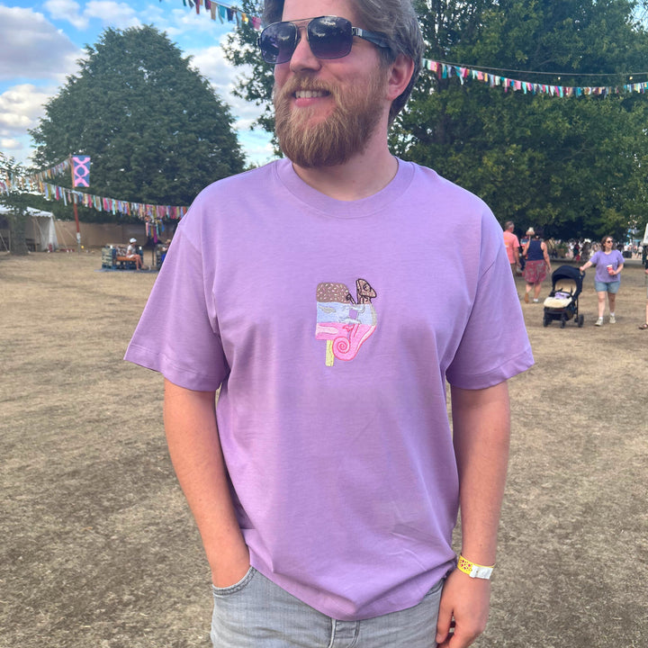 A man wearing a lavender purple t-shirt with a pink and brown graphic on the chest, paired with light gray denim shorts, standing outdoors in a park-like setting with trees and bunting in the background.