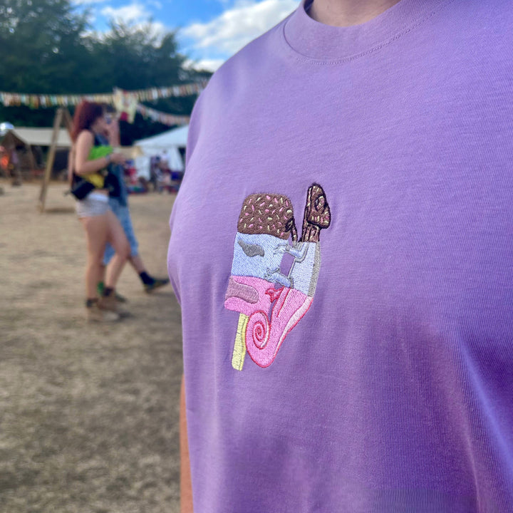 A close-up of a person wearing a lavender purple t-shirt with a colorful embroidered ice cream cone graphic on the chest, set against a blurred outdoor festival background.