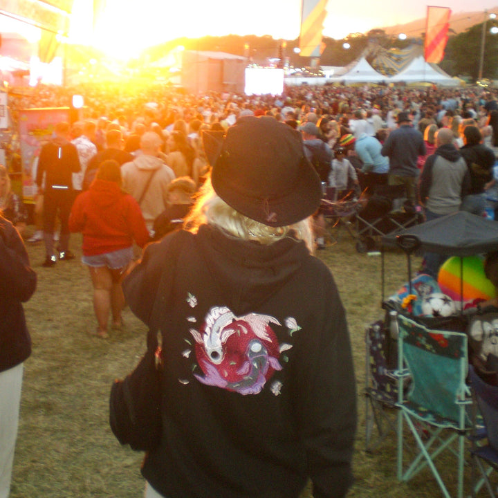 A person standing at an outdoor festival at sunset, viewed from behind wearing a black hoodie with a colorful graphic print on the back and a black cap.