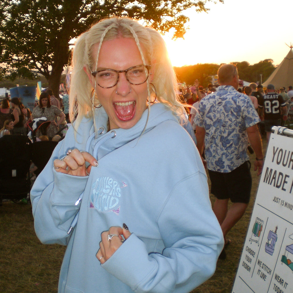 A woman with blonde braids and glasses wearing an oversized light blue hoodie, posing with a wide smile at an outdoor festival during sunset. A sign is visible on the right side of the frame.
