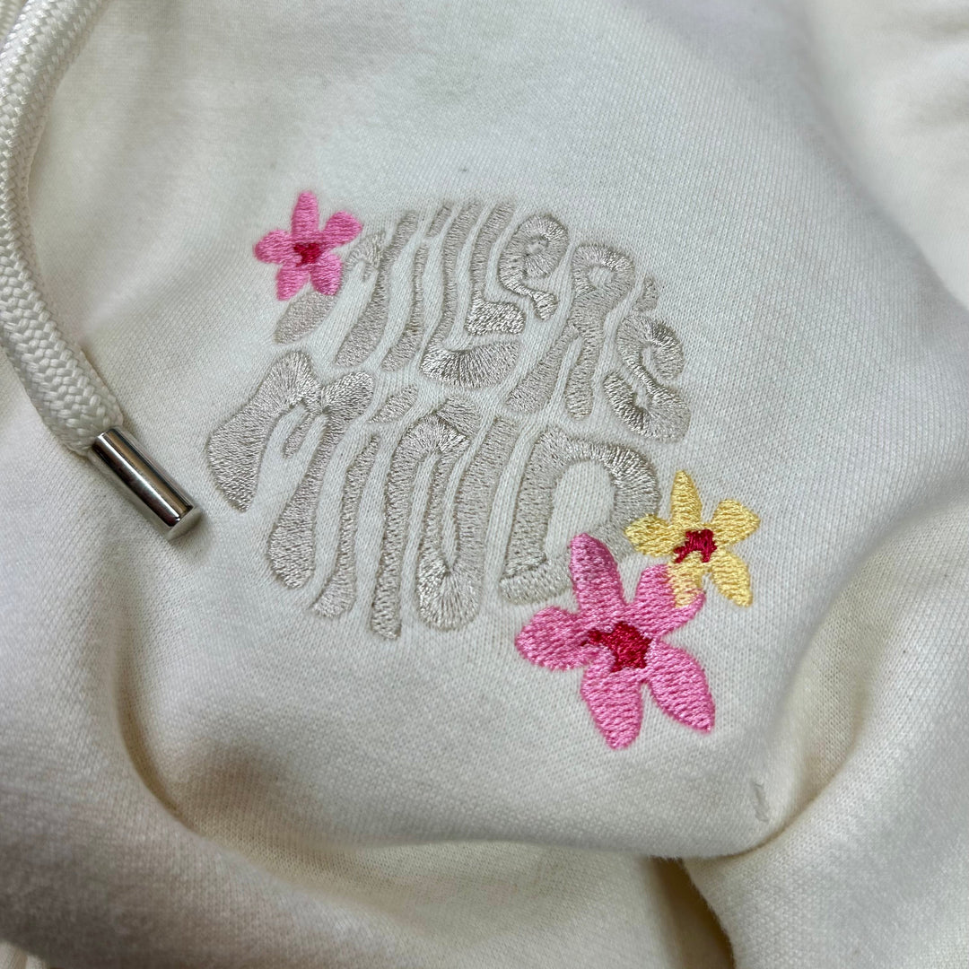 Close-up of a cream-colored hoodie featuring a metallic silver embroidered 'millersmind' design on the chest, accented by two pink flowers and one yellow flower, with a white drawstring visible.