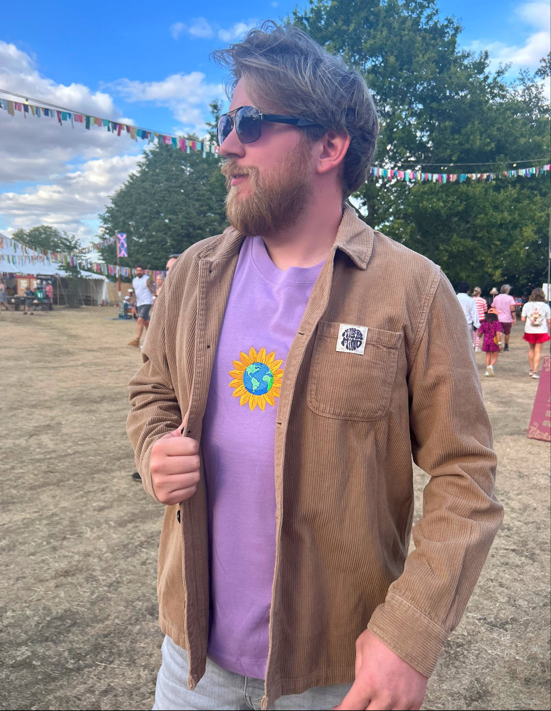 A man wearing a brown corduroy overshirt and a lavender t-shirt with a sunflower around the earth graphic, standing outdoors at what looks like a festival or fairground.