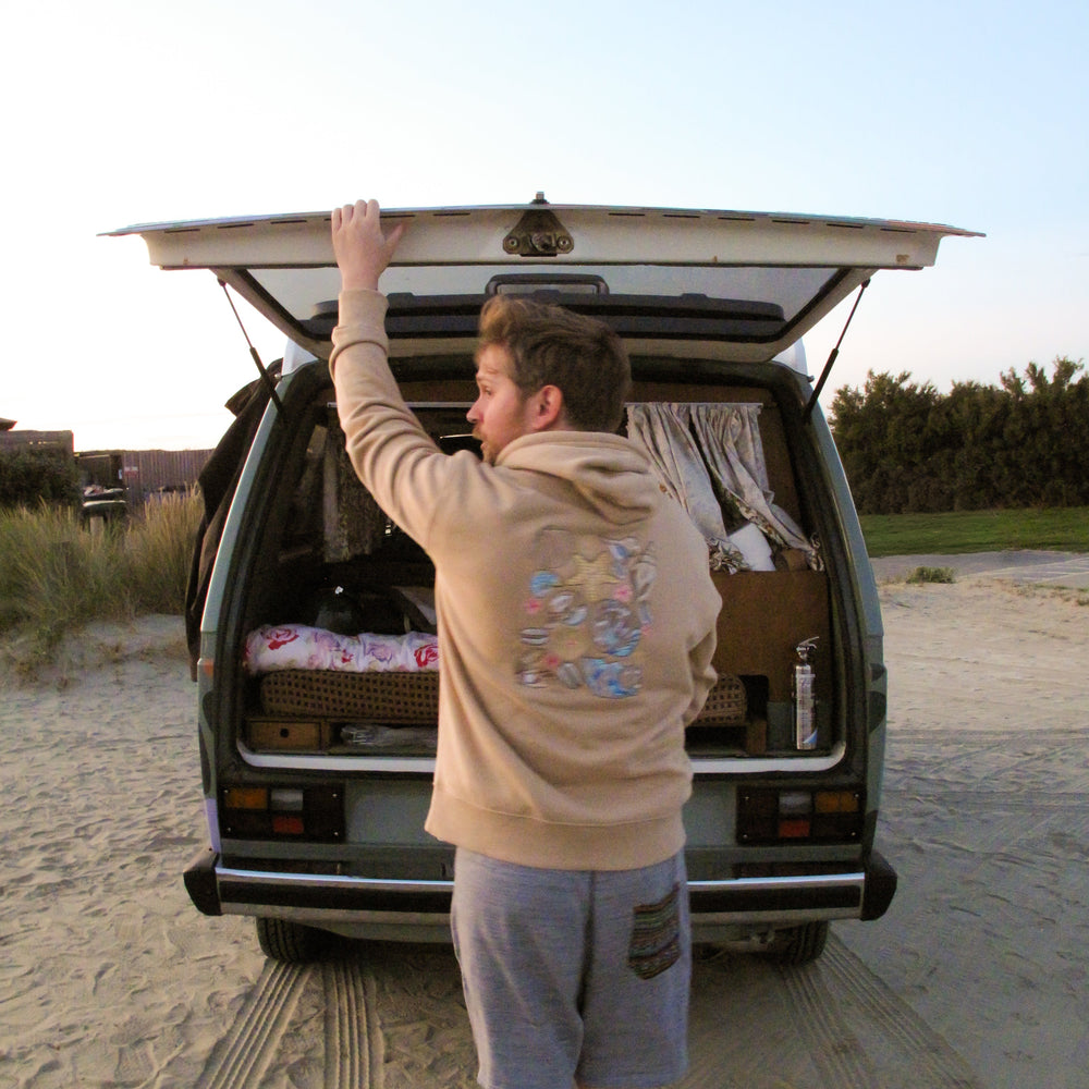 A man in a beige hoodie and gray shorts stands in front of an open camper van on a sandy beach, reaching up to close the tailgate. The interior of the van is styled with bedding and curtains.