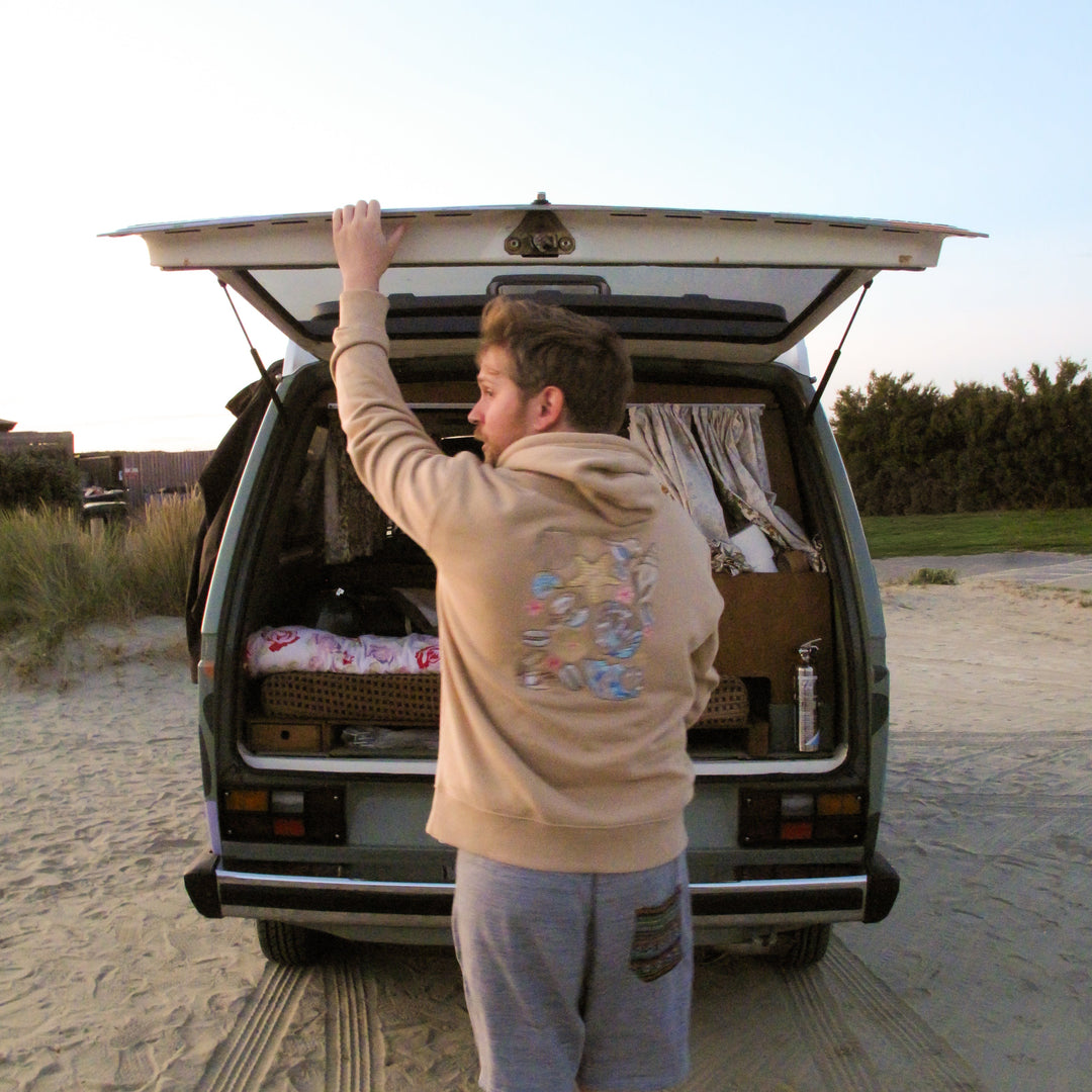 A man in a beige hoodie and gray shorts stands in front of an open camper van on a sandy beach, reaching up to close the tailgate. The interior of the van is styled with bedding and curtains.