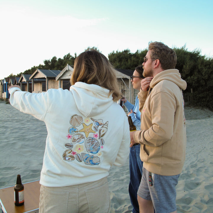 Three people standing on a sandy beach near beach huts, with the person in the foreground wearing a white hoodie featuring a colorful seashell and starfish graphic on the back.