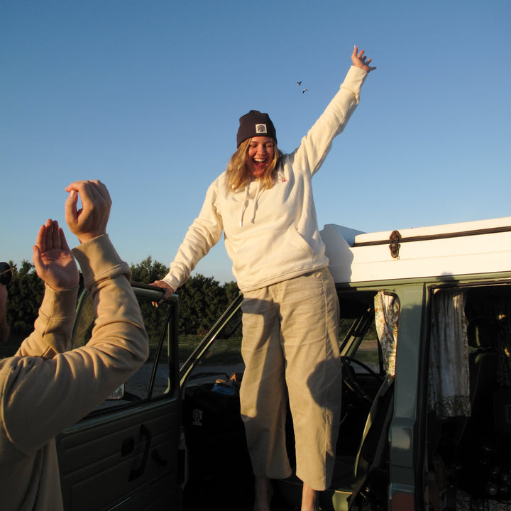 A woman stands barefoot on the open door of a vintage camper van, smiling and waving with one arm raised. She is wearing a cream hoodie, beige cropped pants, and a dark beanie.