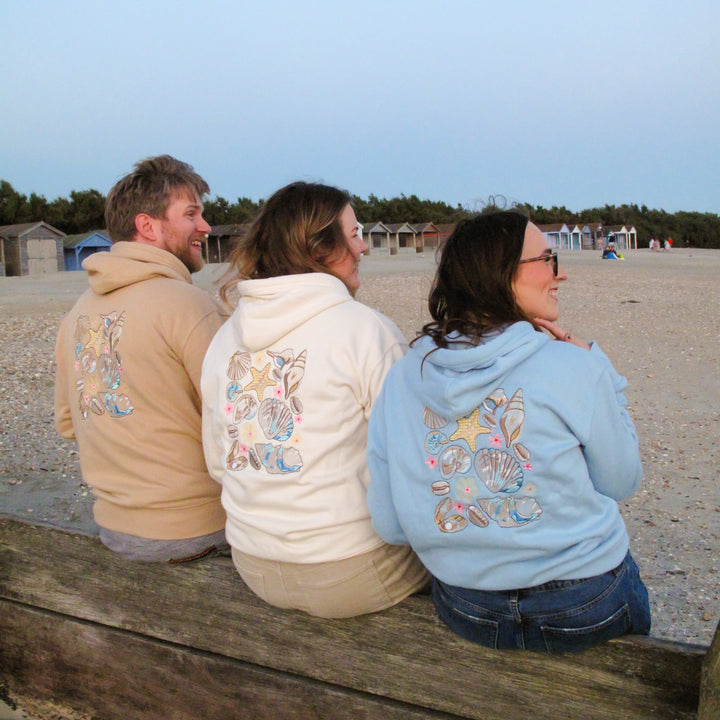 Three people sitting on a wooden bench at a beach, wearing matching hoodies with a large colorful fish graphic on the back. The hoodies are in beige, cream, and light blue colors.