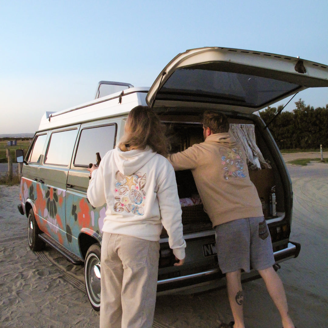 Two people standing at the open trunk of a vintage-style camper van parked outdoors on a sandy surface under a clear blue sky. The van has a multicolor retro pattern on the side.