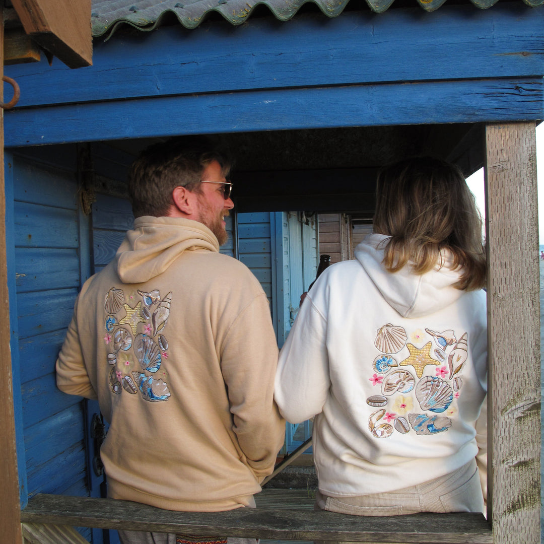 Two people standing on a wooden porch in front of a blue wooden building, wearing hoodies with colorful seashell and star graphics on the back.