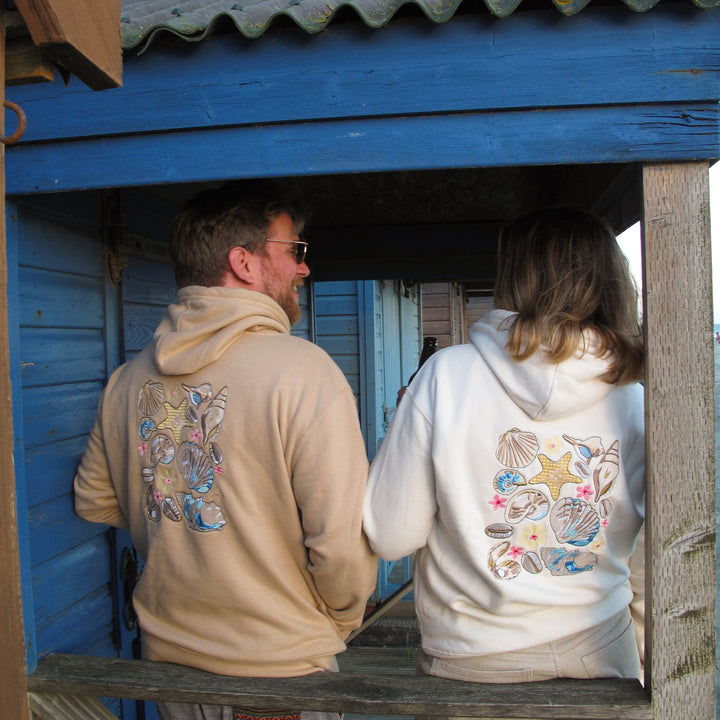 Two people standing on a wooden porch in front of a blue wooden building, wearing hoodies with colorful seashell and star graphics on the back.