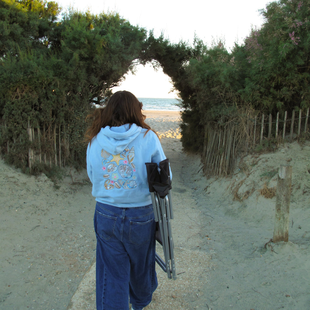 A person viewed from behind walking on a sandy path between dune shrubs toward the ocean, carrying a black folding chair. The person wears a light blue hoodie with a colorful graphic on the back and dark blue jeans.