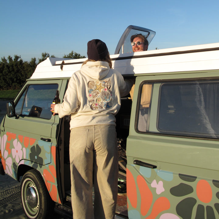 A person in a beige hoodie and sweatpants stands by the open door of a retro-style green van with floral decals, while another person looks out from the elevated window under a clear blue sky.