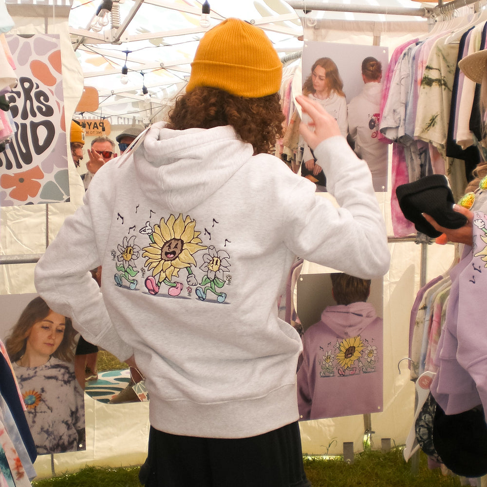 A person standing in a market booth wearing a white hoodie with a colorful sunflower graphic on the back, paired with black shorts and a yellow beanie. Clothing racks and posters are visible in the background under a tent.