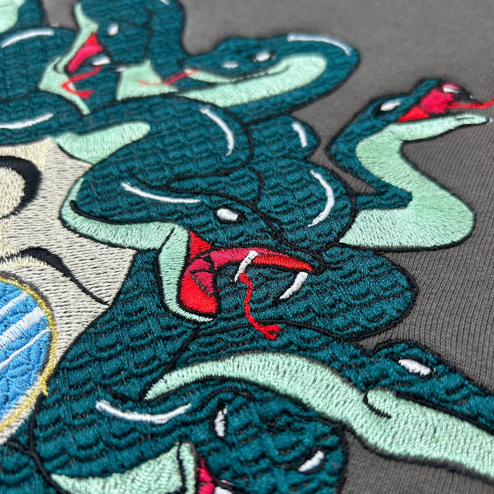 Close-up of detailed embroidery depicting multiple teal and mint green snakes with red tongues on dark fabric, inspired by a modern Medusa theme