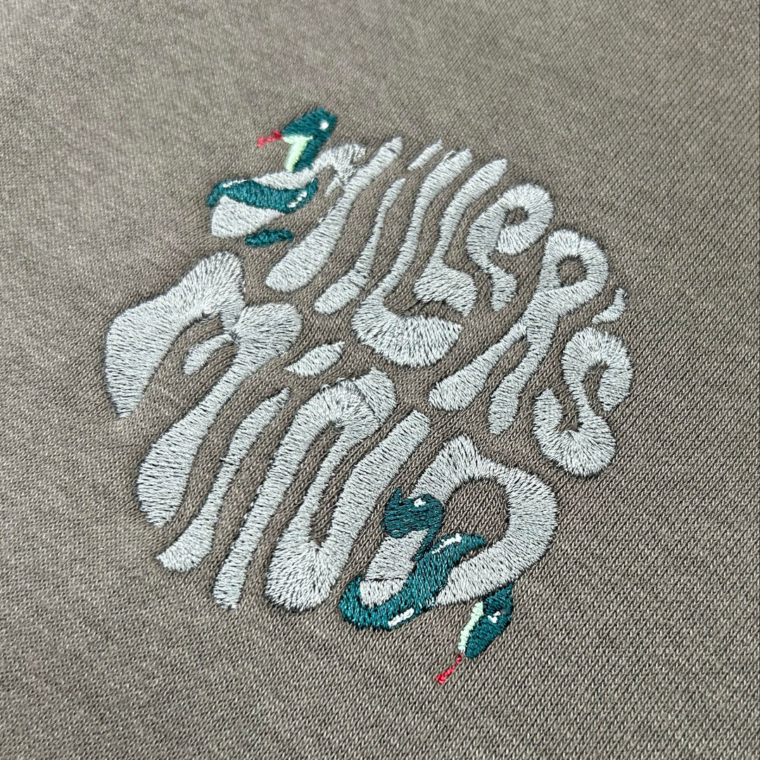 Close-up of detailed embroidery depicting multiple teal and mint green snakes with red tongues on dark fabric, inspired by a modern Medusa theme