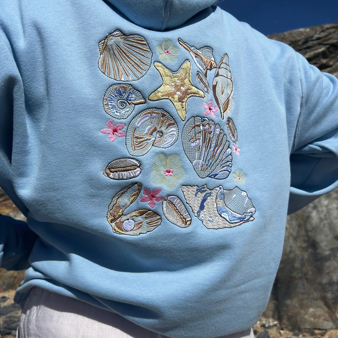Light blue hoodie with embroidered seashells and starfish design against a natural background.