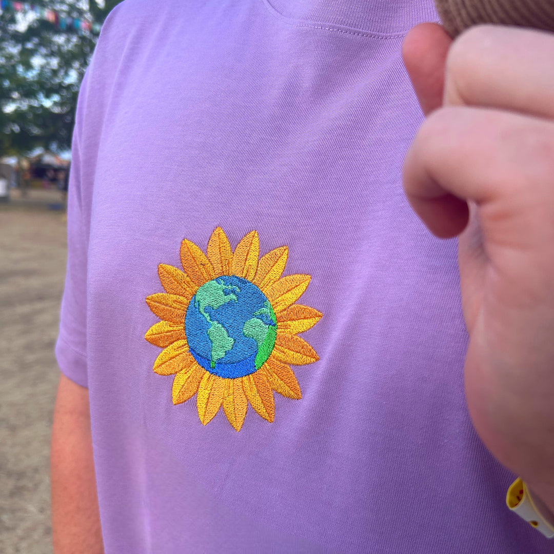 A close-up of a person wearing a lavender purple t-shirt with an embroidered sunflower design featuring a blue and green globe in the center. A hand wearing a brown knit glove is partially visible in the foreground.