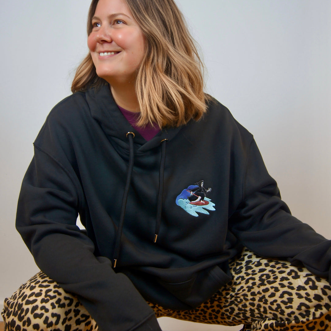 women wearing a black hoodie with a small embroidery of a surfing reaper. All made from MillersMind