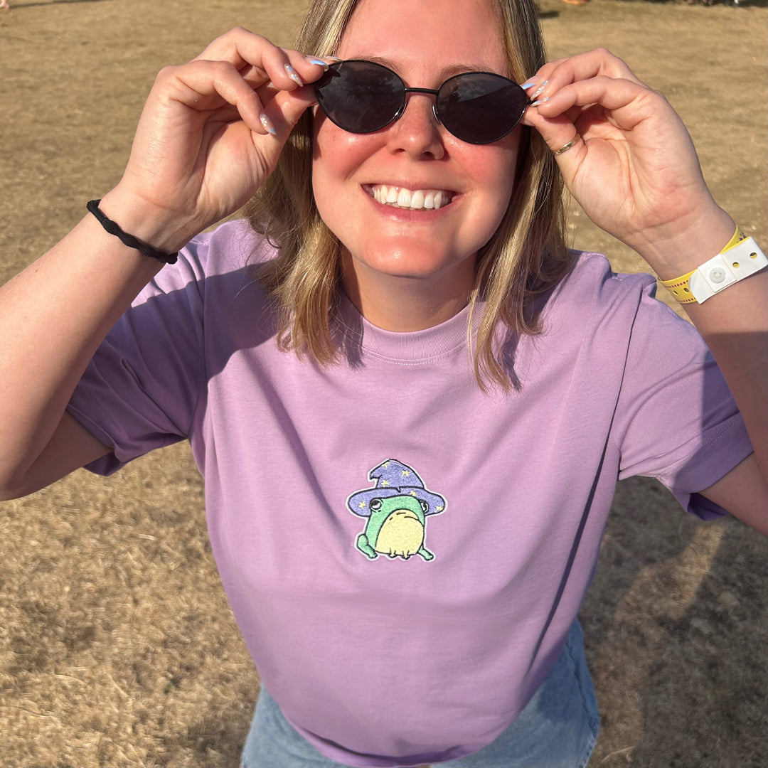 A smiling woman outdoors wearing a lavender-purple t-shirt with a small cartoon frog graphic on the chest, holding her sunglasses up to her eyes.