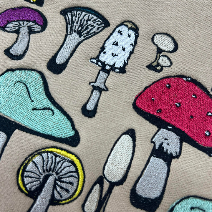 Close-up view of colourful embroidered mushrooms on olive green fabric, featuring detailed designs in shades of yellow, purple, teal, red, and orange.