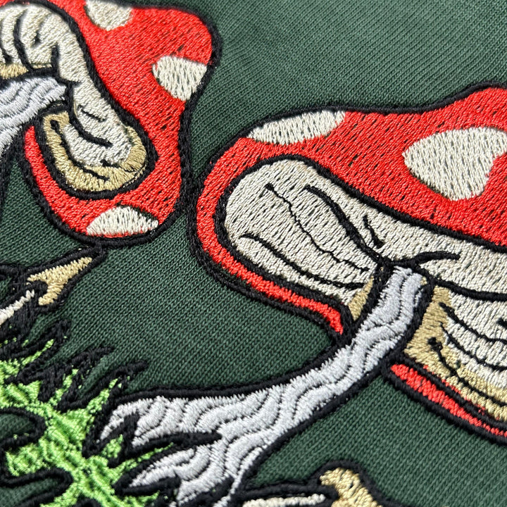 Woman wearing a forest green T-shirt with embroidered red and white mushrooms on the chest, paired with cream-colored pants covered in colourful mushroom prints, kneeling against a plain light background
